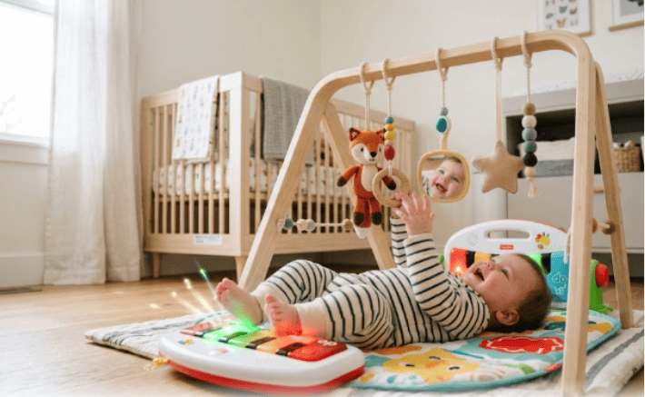 A baby lying on their back kicking a light-up piano at the foot of a wooden baby gym while looking at hanging toys. best baby gyms