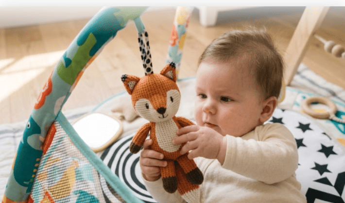 A close-up of a 4-month-old baby lying on a play mat and reaching out to grasp a hanging knit fox sensory toy.best baby gyms